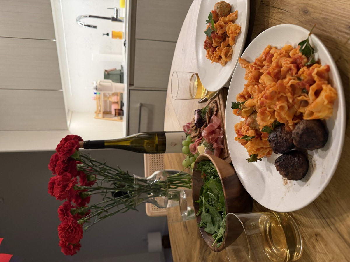 Close-up of pasta plates with meatballs, charcuterie board, wine, and red carnations